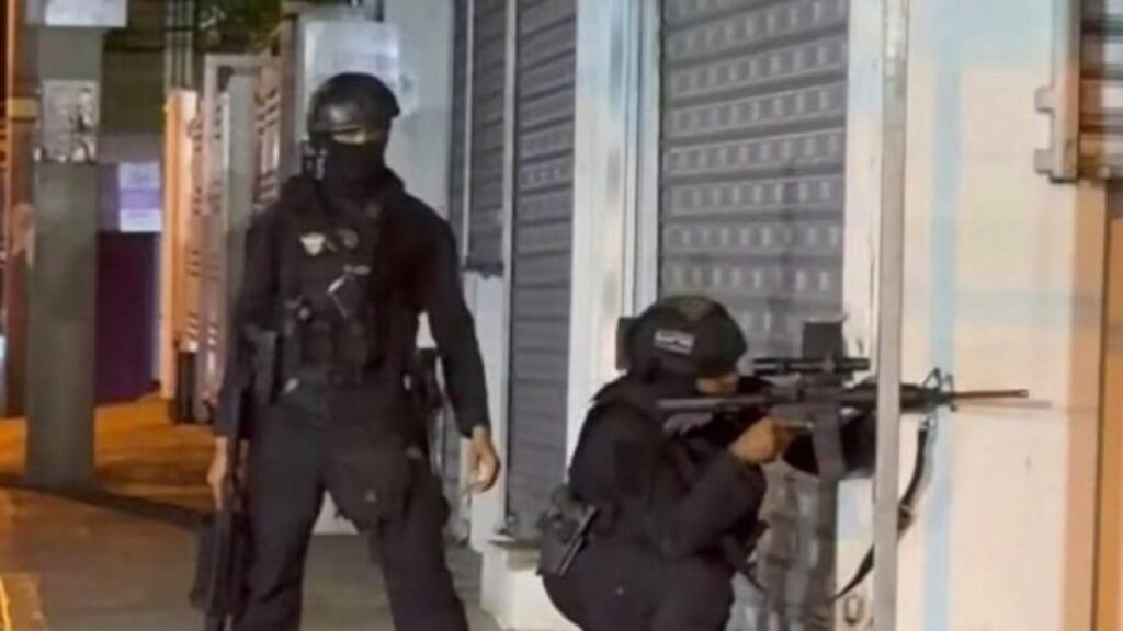 Dos agentes con uniforme negro, casco y pasamontañas, uno de ellos agachado y apuntando con un fusil, junto a comercios cerrados durante la noche en Salcedo